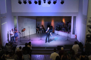 Fotografia de um culto realizado com o ministro no centro do palco acompanhado pela banda